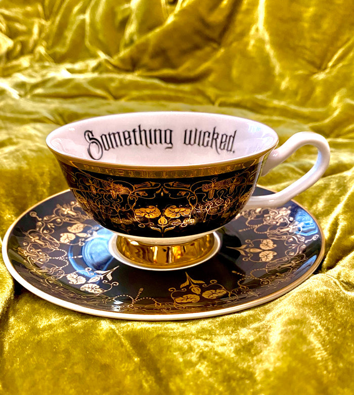 Black and Gold Something Wicked Nouveau Teacup and Saucer positioned on a rich green fabric, featuring the text 'Something wicked' and ornate gold detailing.